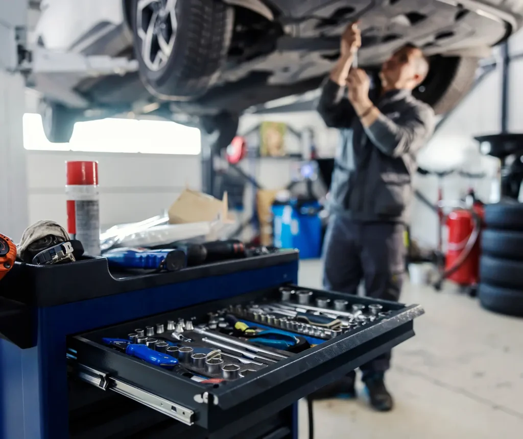 About us, Auto repair shop in Springboro, OH, Springboro Automotive. A mechanic works underneath a lifted car in a garage. In the foreground, a tool cart with an open drawer reveals various tools, including wrenches and screwdrivers. The garage setting includes equipment like cans and stacked tires in the background.