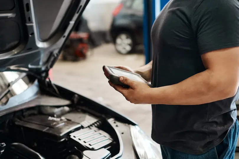 Careers, Auto repair shop in Springboro, OH, Springboro Automotive. A mechanic holding a tablet inspects a car engine in a garage. The cars hood is open, revealing the engine. A blurred vehicle is visible in the background. The mechanic is wearing a dark t-shirt.