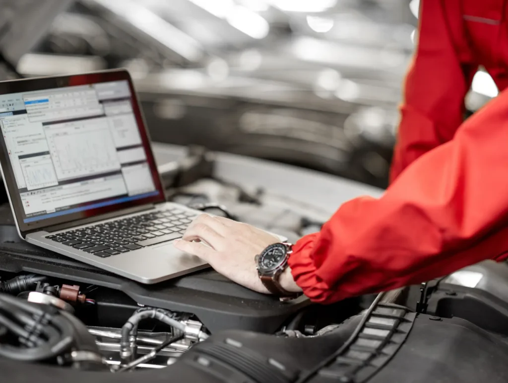 car maintenance near me, Auto repair shop in Springboro, OH, Springboro Automotive. A person in a red jacket uses a laptop on the hood of a car, which is open for maintenance. The laptop displays diagnostic software. The setting appears to be a garage or workshop.