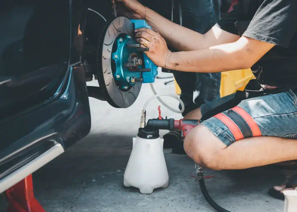 brake repair, Auto repair shop in Springboro, OH, Springboro Automotive. A person in shorts is working on a cars brake system, using a brake bleeder tool. The wheel is off, revealing the blue brake caliper. The setting is outdoors on a concrete surface.