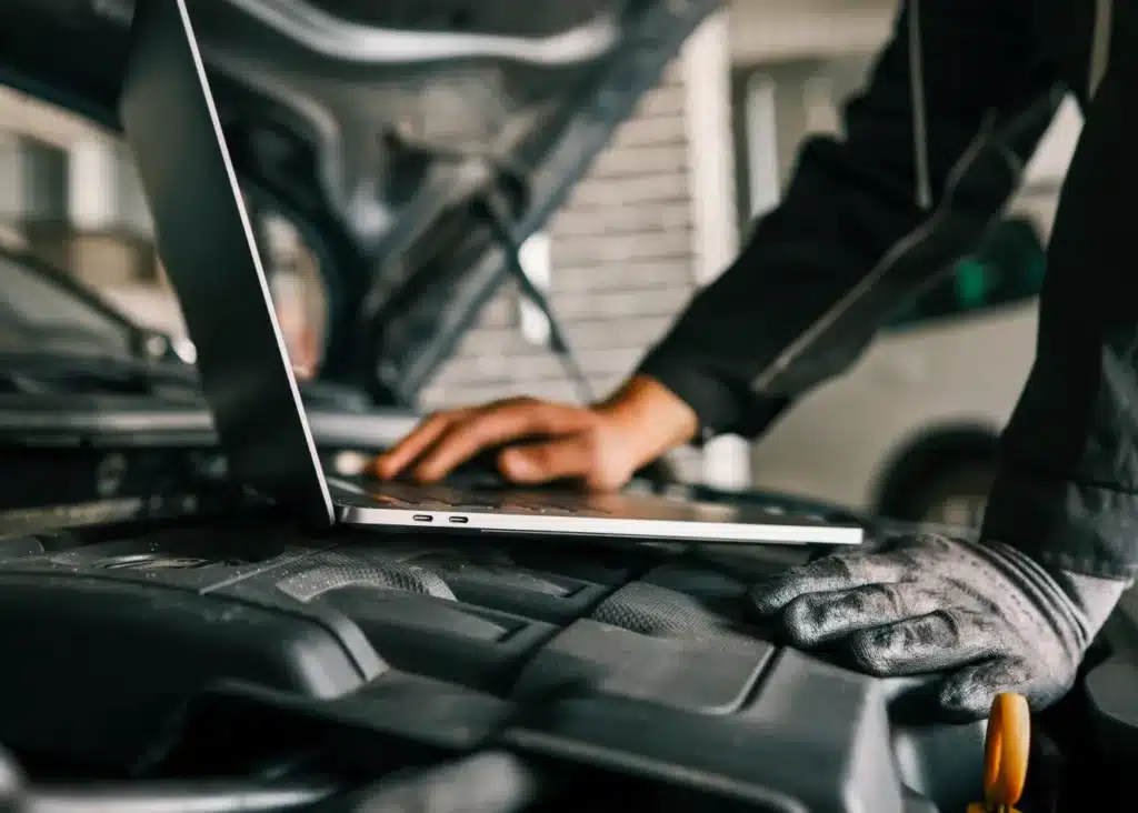 auto engine repair and diagnostics, Auto repair shop in Springboro, OH, Springboro Automotive. A person in work gloves and overalls uses a laptop placed on the open hood of a car, possibly diagnosing or repairing the vehicle. The cars engine is partially visible.