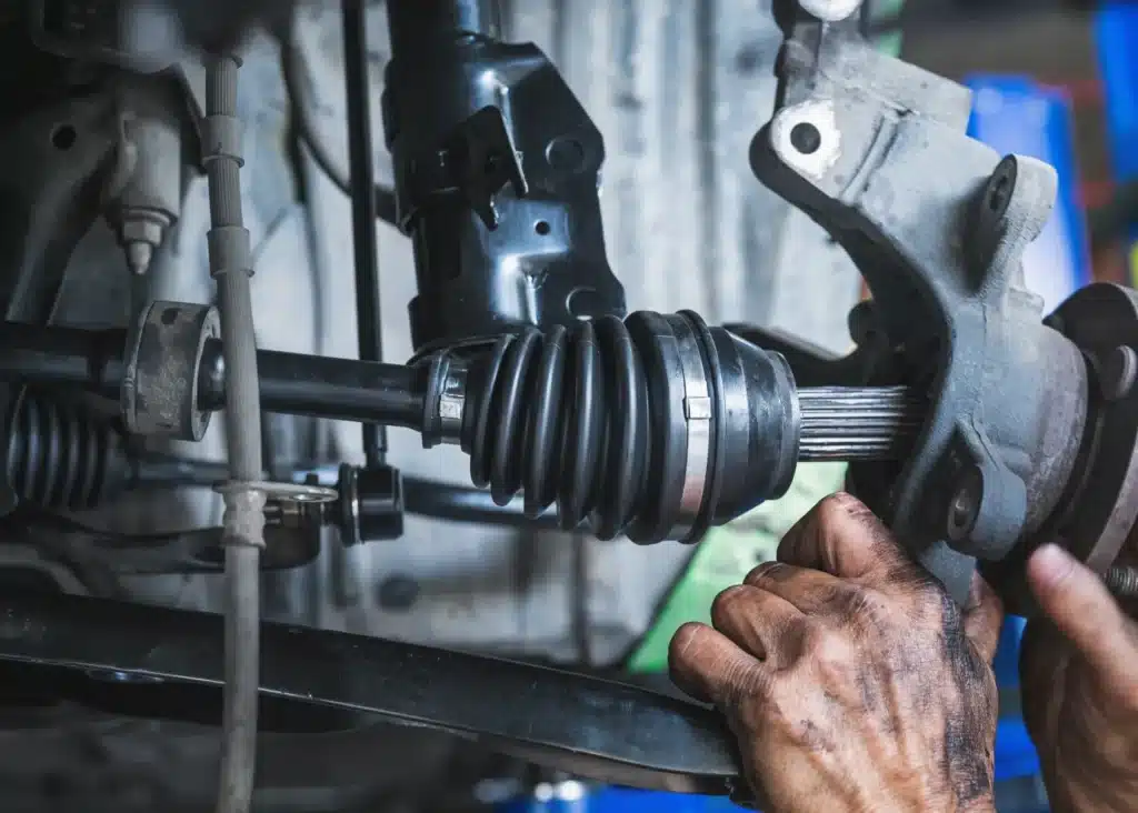 steering and suspension repair, Auto repair shop in Springboro, OH, Springboro Automotive. Close-up of a mechanics hands working on a cars suspension system. The image shows dirty hands adjusting the CV joint and axle, with metal parts and a rubber boot visible. The background is slightly blurred, focusing on the mechanical components.
