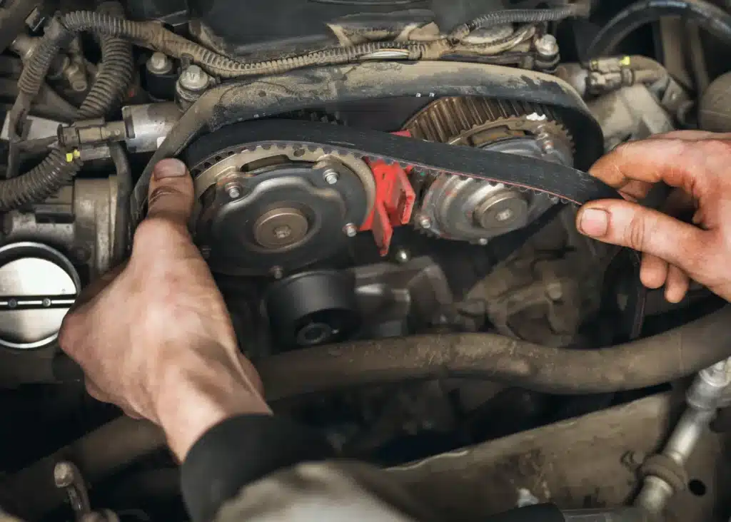 Timing belt replacement, Auto repair shop in Springboro, OH, Springboro Automotive. A persons hands are adjusting a timing belt on the exposed engine of a car. The image shows various mechanical parts, wires, and gears. The setting appears to be in a workshop or garage.