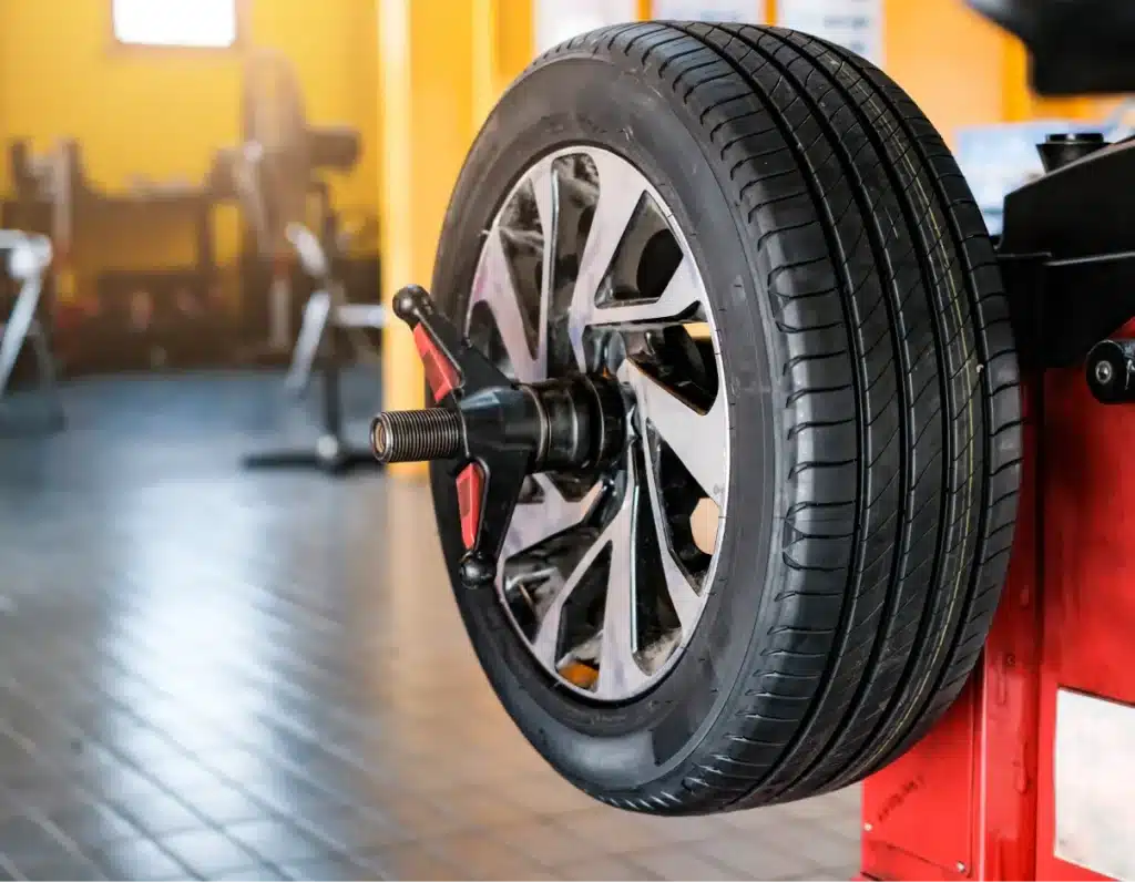 tire services, Auto repair shop in Springboro, OH, Springboro Automotive. A car tire is mounted on a wheel balancing machine in a workshop with a tiled floor. The machine has red and black components, and the background includes yellow walls and blurred workshop equipment.