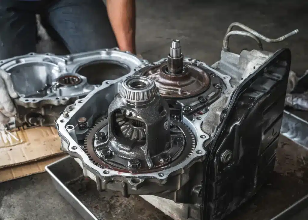 Transmission services, Auto repair shop in Springboro, OH, Springboro Automotive. Close-up of a person inspecting and working on a disassembled car transmission. The gearbox and various metal components, including gears and shafts, are visible. The person is kneeling next to the parts, wearing jeans.