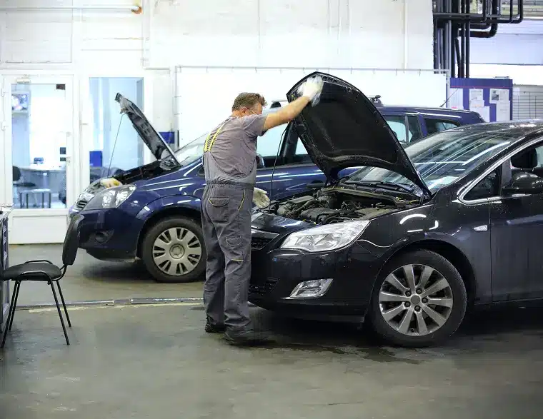 Auto mechanic inspecting the engine of a black car inside a well-lit garage, with another vehicle being serviced in the background.