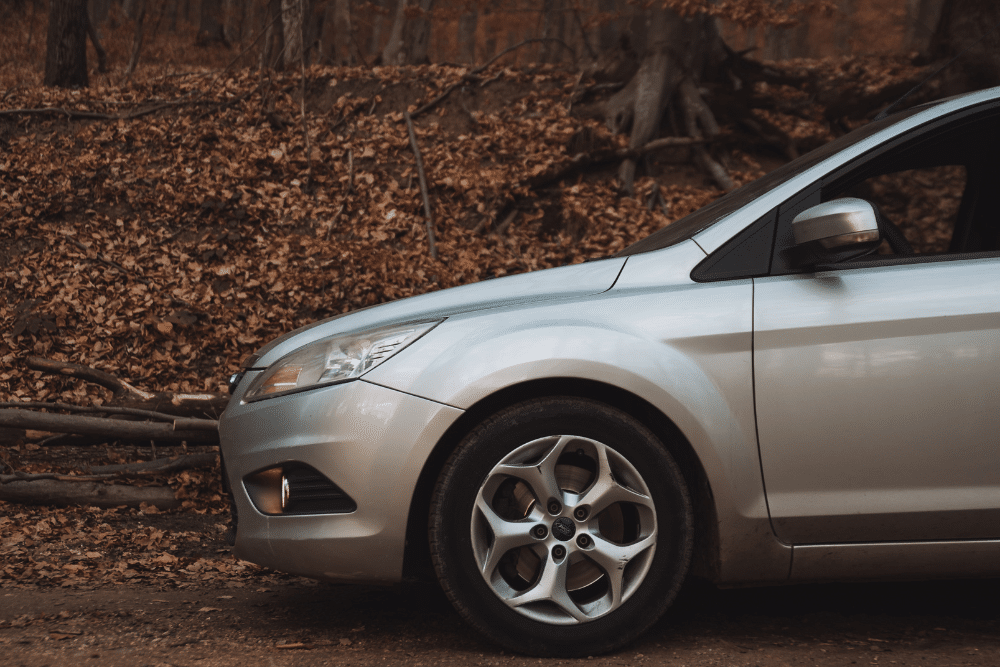 Fall Car Care. Auto repair in Springboro, OH. Springboro Automotive. A silver car parked on a leaf-covered road in a forest during autumn, symbolizing fall car care and preparation for winter driving.
